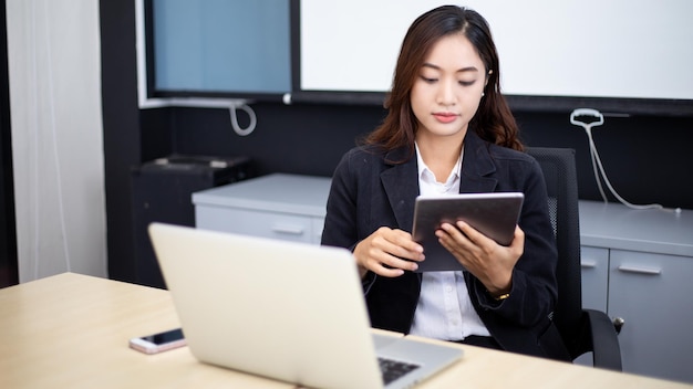 Lächelnde asiatische Geschäftsfrau, die ein Tablet für die Arbeit verwendet, und ein süßes Mädchen, das nach Daten sucht und sich auf ihr Tablet in ihrem Büro konzentriert