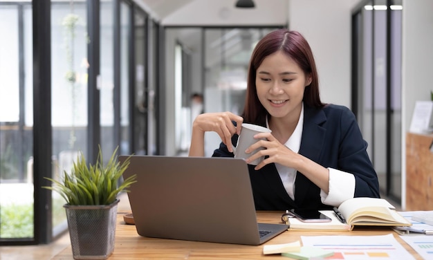 Lächelnde asiatische Geschäftsfrau, die eine Kaffeetasse und einen Laptop im Büro hält