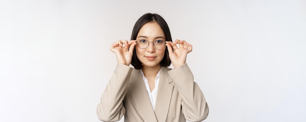 Lächelnde asiatische Geschäftsfrau, die eine neue Brille mit Brille versucht, die im Anzug auf weißem Hintergrund steht