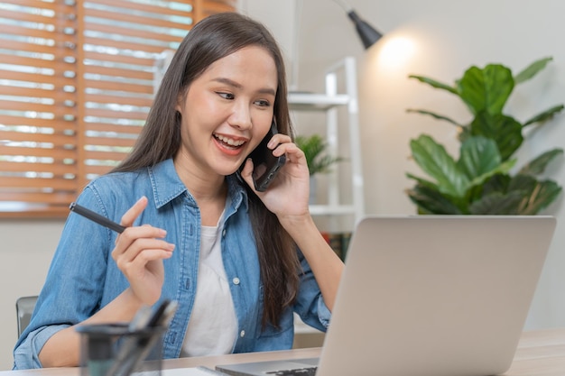 Lächelnde asiatische junge professionelle geschäftsfrau des unternehmers, die auf dem schreibtisch auf dem schreibtisch das smartphone hält, das mit dem kunden spricht, während sie im büro zu hause sitzt