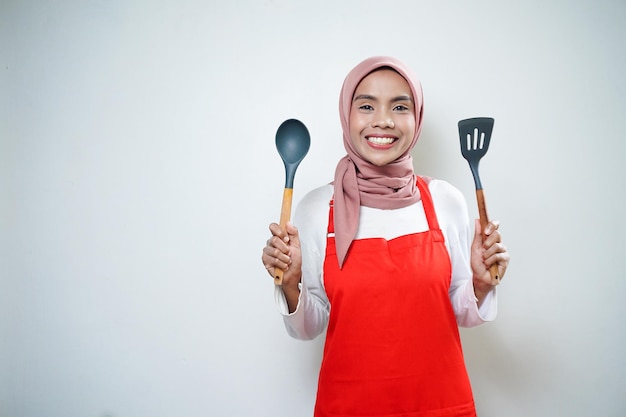 Lächelnde asiatische muslimische Frau in roter Schürze mit Suppenlöffel und Pfannenwender Kochgeschirr Kochkonzept