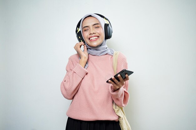 Lächelnde asiatische muslimische Studentin in rosafarbenem Pullover mit Tasche und Handy, die Musik hört