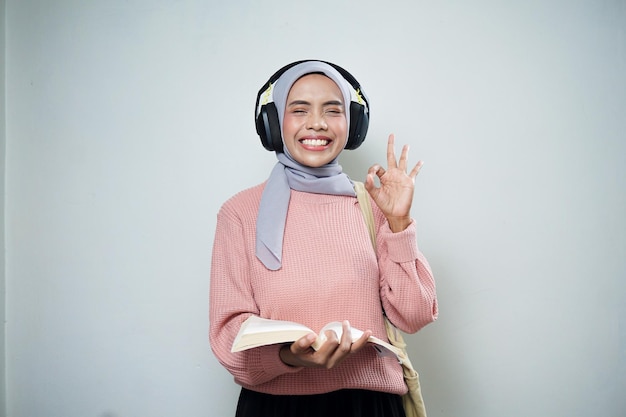 Lächelnde asiatische muslimische Studentin in rosafarbenem Pullover mit Tasche, während sie Musik hört und Buch liest