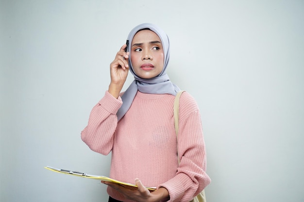 Lächelnde asiatische muslimische Studentin in rosafarbenem Pullover mit Taschenhaltebrett und leerem Brett