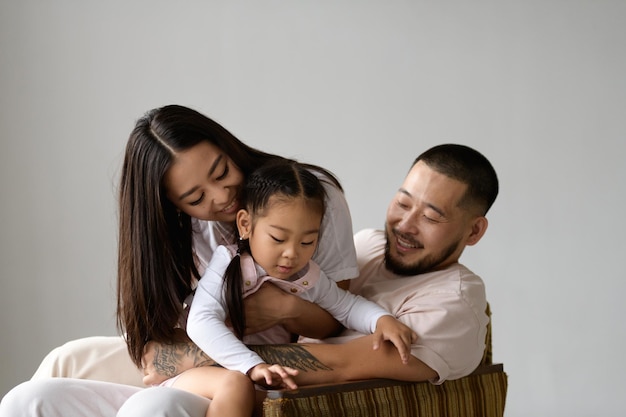 Lächelnde asiatische Mutter umarmt ihre Kleinkindtochter in der Nähe ihres Mannes auf einem Sessel, isoliert auf grauem Hintergrund