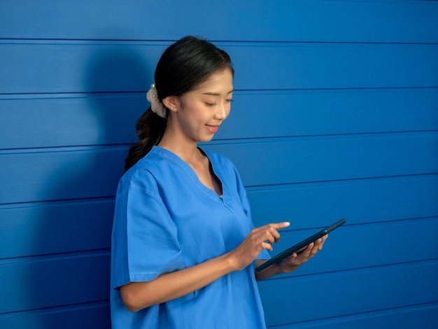 Lächelnde asiatische Ärztin oder Krankenschwester in blauem Peeling-Anzug mit Smartphone, während sie auf blauem Hintergrund steht und auf den Bildschirm blickt, der Online-Chat-Beratung des Patienten-Telemedizin-Konzepts arbeitet