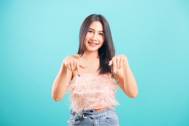 Lächelnde asiatische schöne Frau mit dem Zeigefinger nach unten und Blick auf die Kamera auf blauem Hintergrund, mit Kopierraum für Text