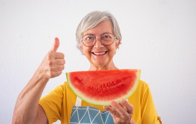 Lächelnde attraktive alte Seniorin, die ein Stück frische Wassermelone hält und mit dem Daumen nach oben in die Kamera schaut, gesundes Ernährungslebensstilkonzept