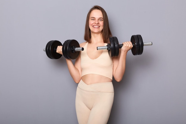 Lächelnde, attraktive, charmante, positive Frau im Top, die Langhanteln in den Händen hält, isoliert auf grauem Hintergrund, beim Training für die Arme und beim Sporttraining