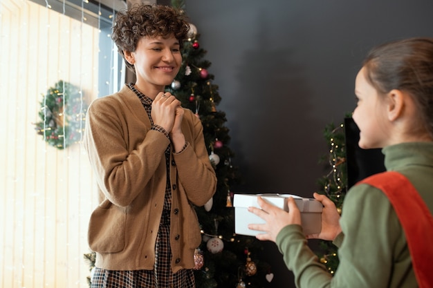 Lächelnde attraktive Mutter in Strickjacke mit gefalteten Händen, die zu Weihnachten eine Geschenkbox von der Tochter bekommt