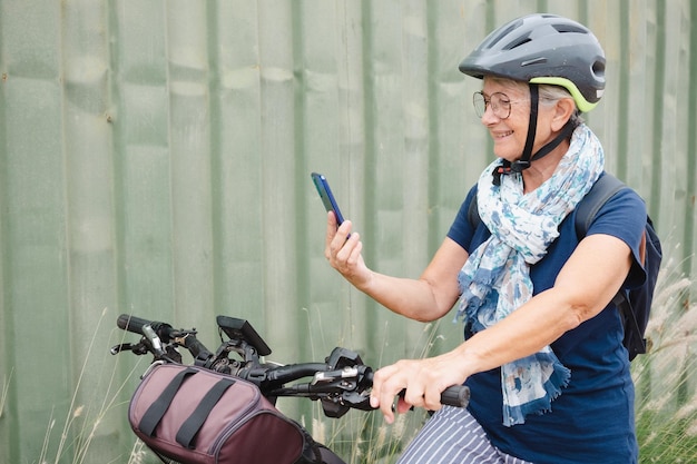 Lächelnde, attraktive Seniorin mit Helm und Rucksack, die Sportaktivitäten mit Elektrofahrrad im Freien genießt, ältere Frau mit Telefon auf grünem Hintergrund