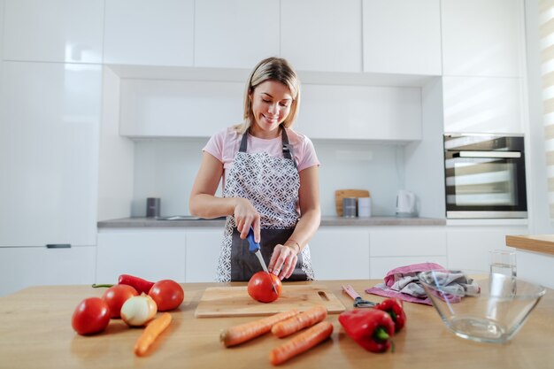 Lächelnde attraktive würdige kaukasische blonde Frau in der Schürze, die Tomate schneidet, während in der Küche steht. Auf der Küchentheke stehen Karotten, Tomaten und Paprika.