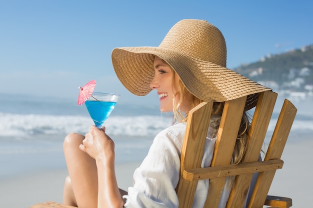 Lächelnde blonde Entspannung im Klappstuhl durch das Meer, das Cocktail hält