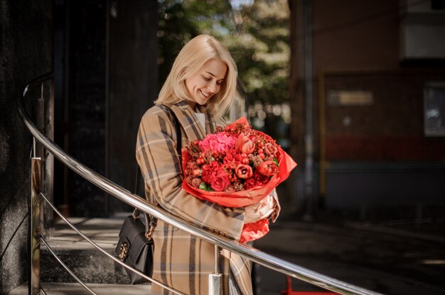 Lächelnde blonde Frau im karierten Mantel mit einem großen roten Blumenstrauß