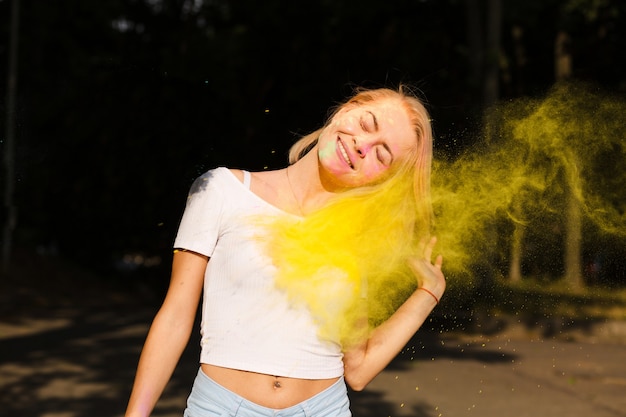 Lächelnde blonde Frau im weißen T-Shirt mit Farbe Holi explodiert in der Nähe ihres Gesichts