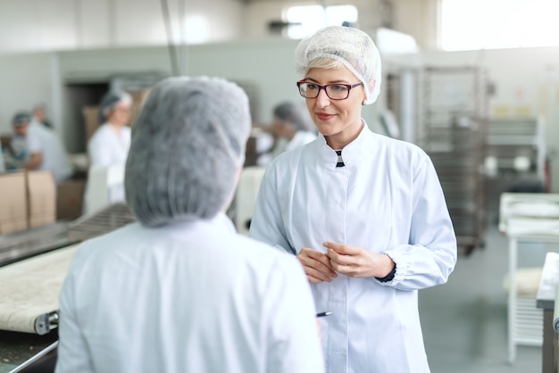 Lächelnde blonde Frau in der weißen sterilen Uniform und mit den Brillen, die mit ihrem Chef sprechen, während sie in der Nahrungspflanze stehen.