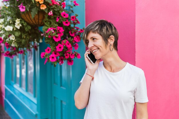 Lächelnde blonde Frau in Freizeitkleidung telefoniert inmitten leuchtend rosa und blauer Häuser in Irland, die mit bunten Pflanzen geschmückt sind