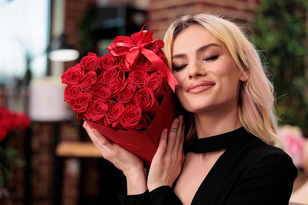 Lächelnde blonde Frau mit roten Rosen aus nächster Nähe, Valentinstag-Luxusgeschenk. Attraktives Mädchen mit Blumenstrauß in herzförmiger Schachtel, teures romantisches Geschenk zum Jubiläum