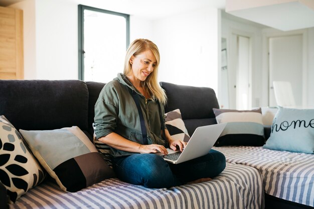 Lächelnde blonde Frau mittleren Alters mit einem Laptop auf dem Sofa eines Wohnzimmers