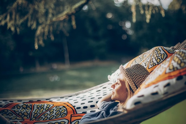 Lächelnde blonde Hipster-Frau, die im Freien auf einer Hängematte schläft Schönes Mädchen in stylischer Jeanskleidung, das sich auf der Hängematte im Park in der Nähe eines See-Camping-Konzepts entspannt