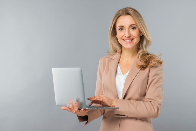 Foto lächelnde blonde junge geschäftsfrau, die in der hand laptop gegen grauen hintergrund verwendet