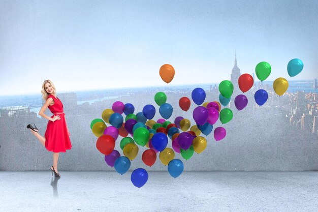 Foto lächelnde blonde stehende hände auf den hüften gegen viele bunte luftballons im raum mit stadt an der wand