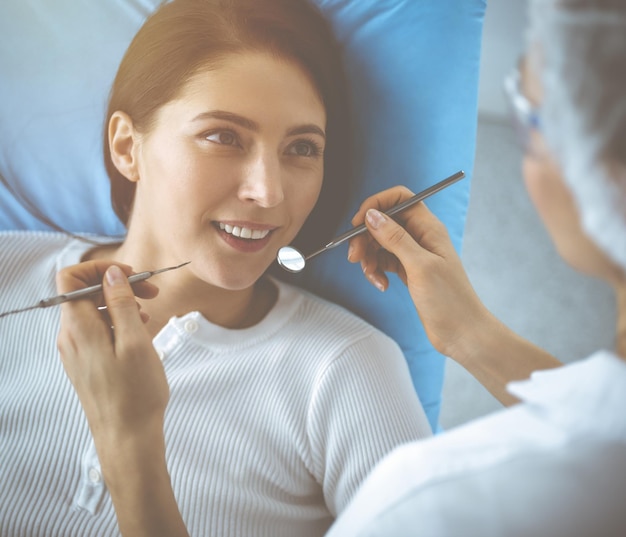 Lächelnde brünette Frau, die vom Zahnarzt in der Zahnklinik untersucht wird. Hände eines Arztes, der zahnärztliche Instrumente in der Nähe des Mundes des Patienten hält. Gesunde Zähne und Medizinkonzept.