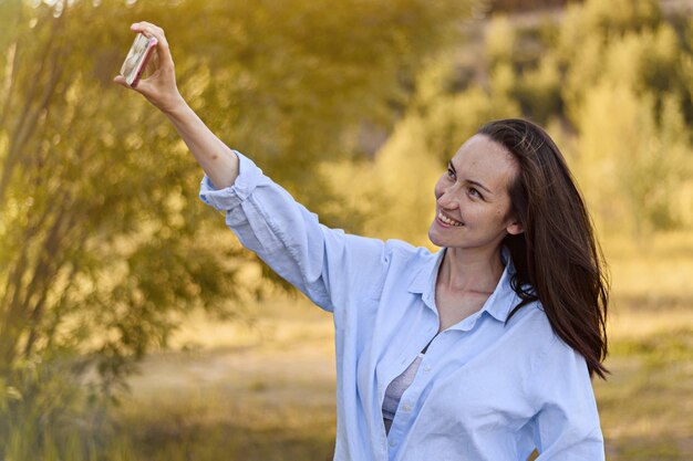 Lächelnde brünette Frau im blauen Hemd nimmt selfie auf Smartphone