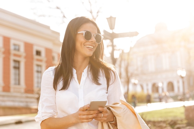 Lächelnde brünette Frau im Hemd und in der Sonnenbrille, die Smartphone halten
