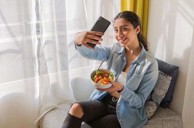 Lächelnde brünette junge Frau, die auf einem Stuhl sitzt und ein selfie mit ihrem Salat in ihrem Zimmer nimmt