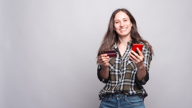 Lächelnde charmante junge Frau, die Smartphone und Kreditkarte über weißer Wand hält