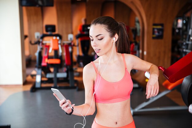 Lächelnde charmante junge Sportlerin in Kopfhörern mit Smartphone und Musikhören im Fitnessstudio