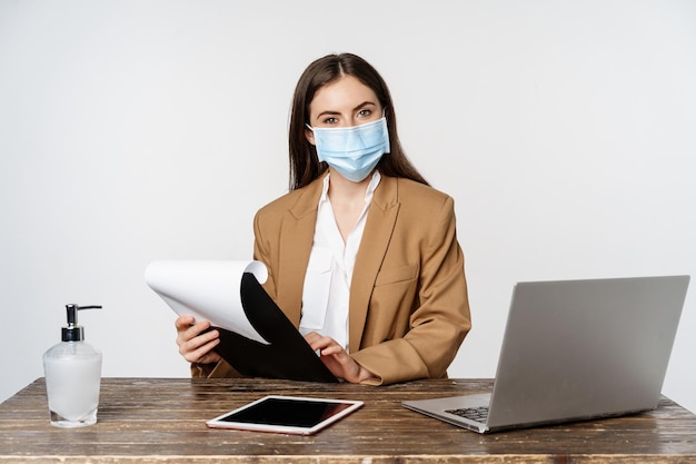 Lächelnde Chefin, weibliche Büroangestellte am Arbeitsplatz, trägt Gesichtsmaske, arbeitet mit Papieren und Dokumenten an ihrem Schreibtisch, weißer Hintergrund