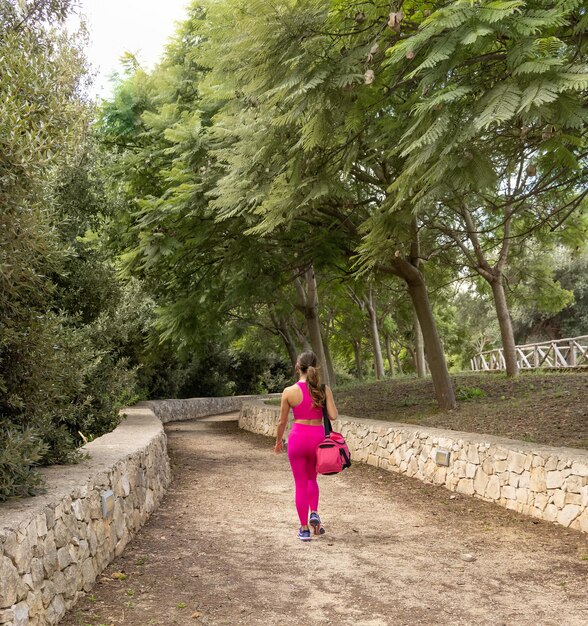 Lächelnde dunkelhaarige Frau gekleidete Sportkleidung, die mit Sporttasche in einem Park spaziert, Rückansicht. Junge kaukasische Frauen in der rosa Kleidung im Freien. Sport und gesundes Lifestyle-Konzept