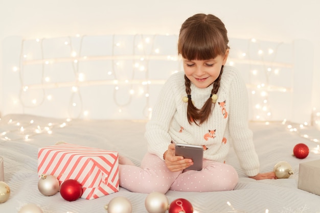 Lächelnde dunkelhaarige Frau mit Zöpfen, die einen weißen Pullover mit dem Handy trägt, Cartoons ansieht oder Spiele spielt, während sie auf dem Bett mit Weihnachtsdekoration sitzt.