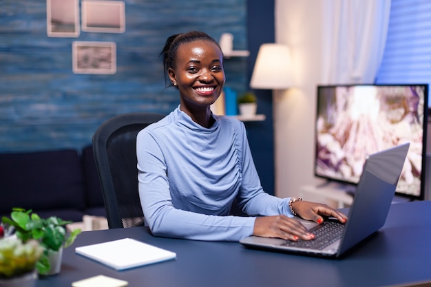 Lächelnde dunkelhäutige Frau lächelt in die Kamera, die am Schreibtisch sitzt und spät in der Nacht vom Home Office aus arbeitet. Schwarzer Freiberufler, der mit einer virtuellen Online-Konferenz im Remote-Team-Chat arbeitet.