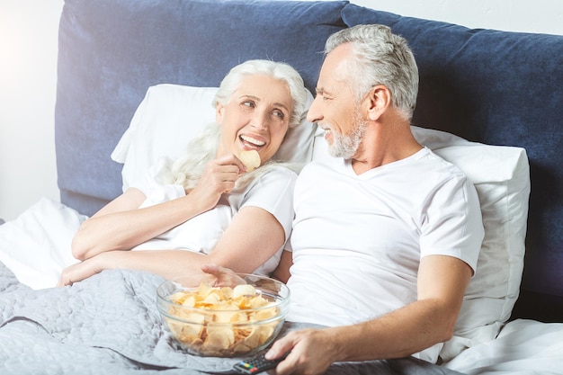 Lächelnde Ehefrau und Ehemann, die fernsehen und Chips im Bett essen