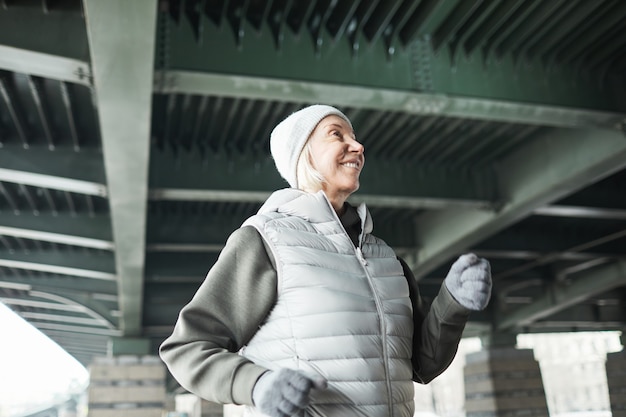 Lächelnde energische ältere kaukasische Frau mit Hut und Handschuhen, die beim Training im Winter allein unter der Brücke joggt