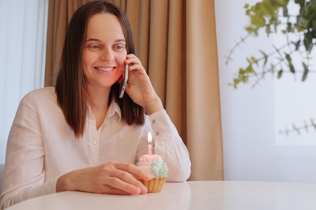 Lächelnde, entzückte junge erwachsene kaukasische Frau in weißem Hemd, die ihren Geburtstag feiert, mit Kuchen und Kerze am Tisch sitzt und telefoniert und Glückwünsche hört