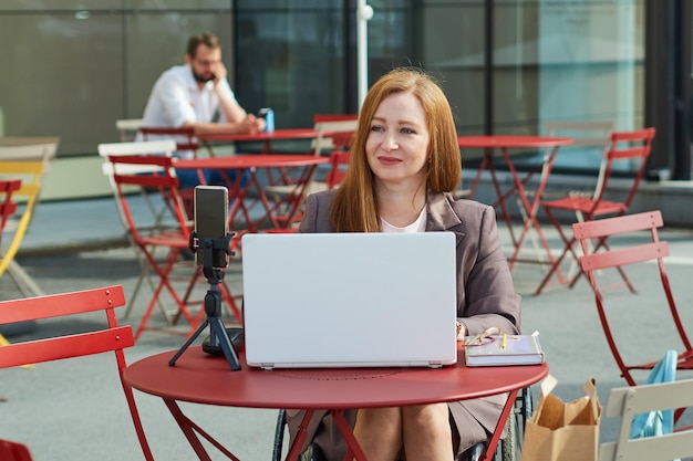 Lächelnde erwachsene Frau, die einen Rollstuhl benutzt und remote am Laptop im Café im Freien arbeitet