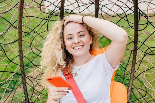 Foto lächelnde europäische frau mit breitem lächeln, das gelockte haar, gekleidet im zufälligen weißen t-shirt, benutzt modernes elektronisches gerät für das surfen von sozialen netzwerken