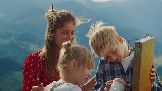 Lächelnde Familie schafft Kunstwerke in windigen Bergen. Niedliche Mutter und Kinder malen im Sommerurlaub auf Staffelei. Positive Frau mit Kindern zeichnet draußen ein Bild der Natur. Kunstunterrichtskonzept