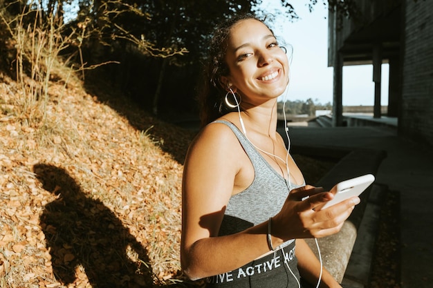 Lächelnde, fitte afrikanische arabische Frau in Tank-Top und Leggings benutzt sein Telefon, während sie bei einem Sonnenuntergang im Freien sitzt, im Freien im Stadion Yoga praktiziert, um sich nach einem stressigen Tag zu entspannen, mit Kopierraum