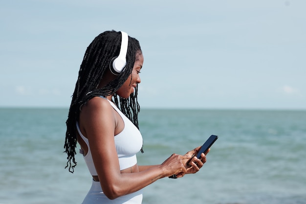 Lächelnde, fitte junge Frau mit Kopfhörern, die Musik für ihr Training über die Anwendung auf dem Smartphone auswählt