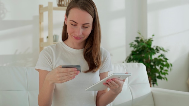 Lächelnde Frau auf der Couch mit einem Tablet und einer Bankkarte macht Online-Einkäufe zu Hause im Wohnzimmer