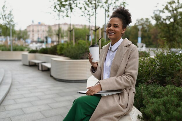 Lächelnde Frau des Porträtafroamerikaners mit Laptop und Tasse Kaffee außerhalb des Bürokonzepts von