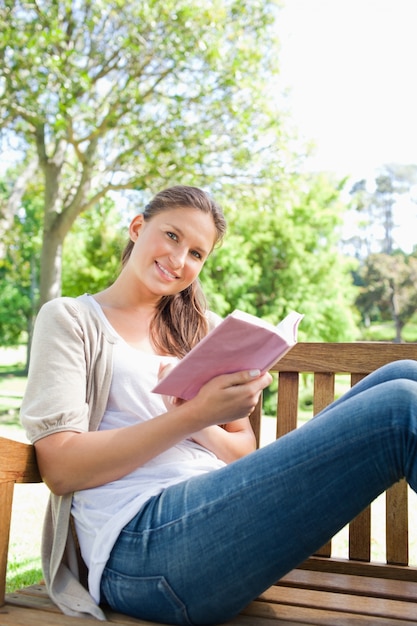 Lächelnde Frau, die auf einer Bank im Park mit einem Buch sitzt