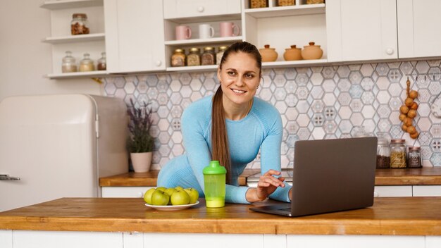 Lächelnde Frau, die Computer im modernen Kücheninnenraum verwendet. Kochen und gesundes Lifestyle-Konzept. Eine Frau schaut in die Kamera und lächelt