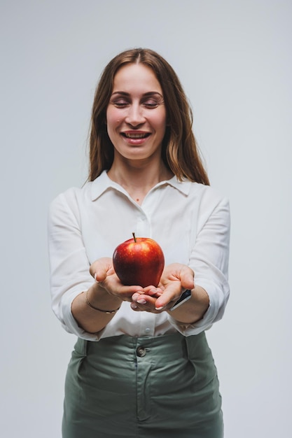 Lächelnde Frau, die einen roten Apfel hält Schöne Brünette in einem weißen Hemd Gesunde Pflanzennahrung und Vitamine Weißer Hintergrund