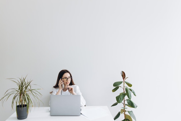 Lächelnde Frau, die mit ihrem Smartphone spricht, während sie mit dem Computer im modernen Startup-Büro arbeitet